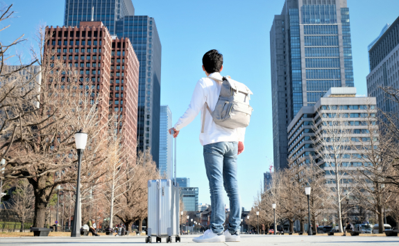 A man stands holding a suitcase. Around him are tall buildings and he is looking up at them in awe